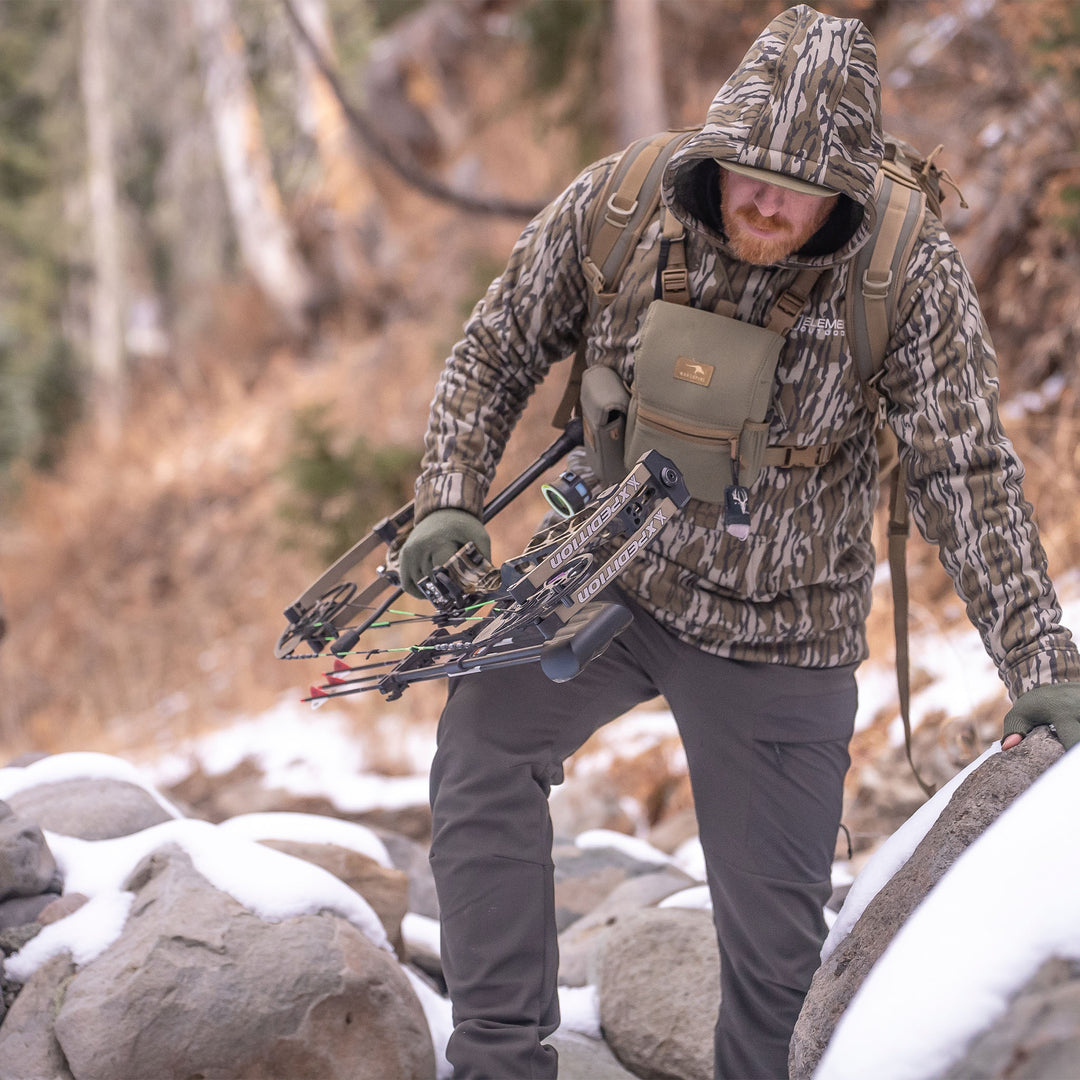 Hunter climbing a hillside wearing a mountaineer hoodie#color_original-bottomland