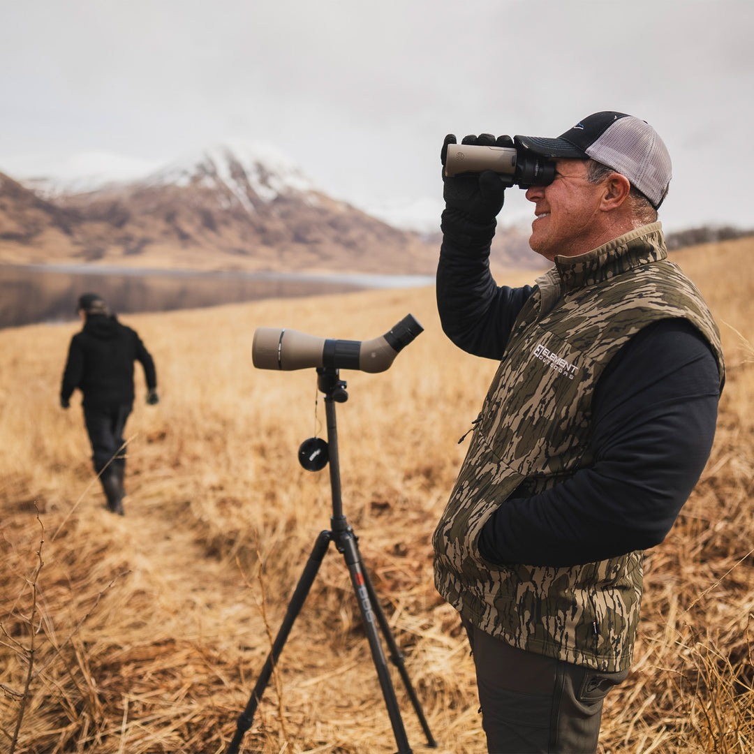 Man looking through Binos in Alaska wearing a Peak Vest#color_original-bottomland